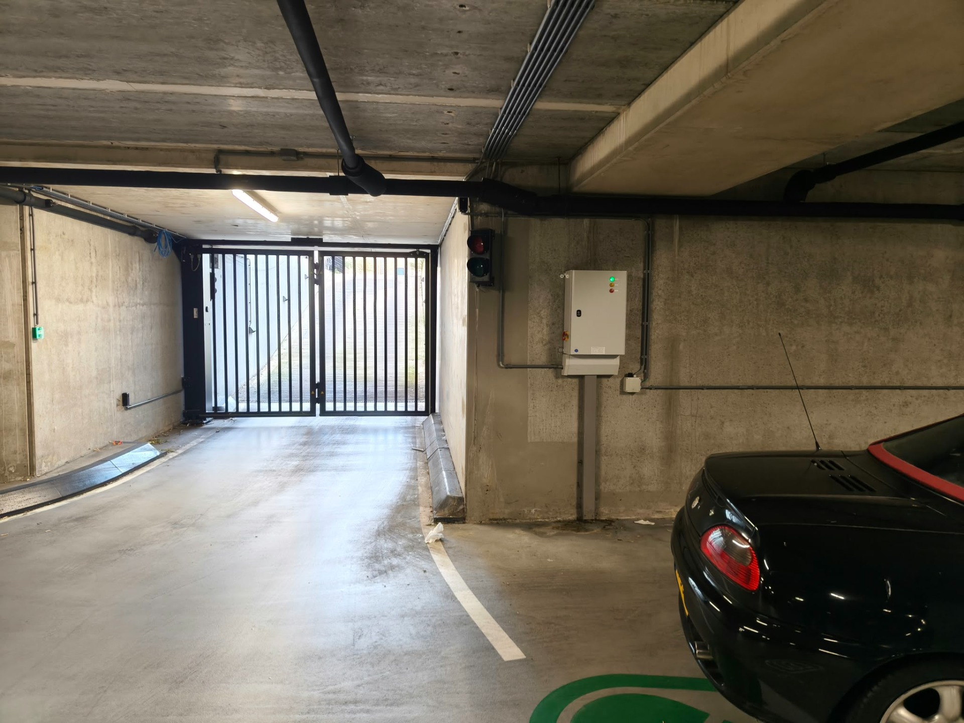Ondergrondse parkeergarage met betonnen wanden, metalen poort, schakelkast en een geparkeerde zwarte auto rechts.
