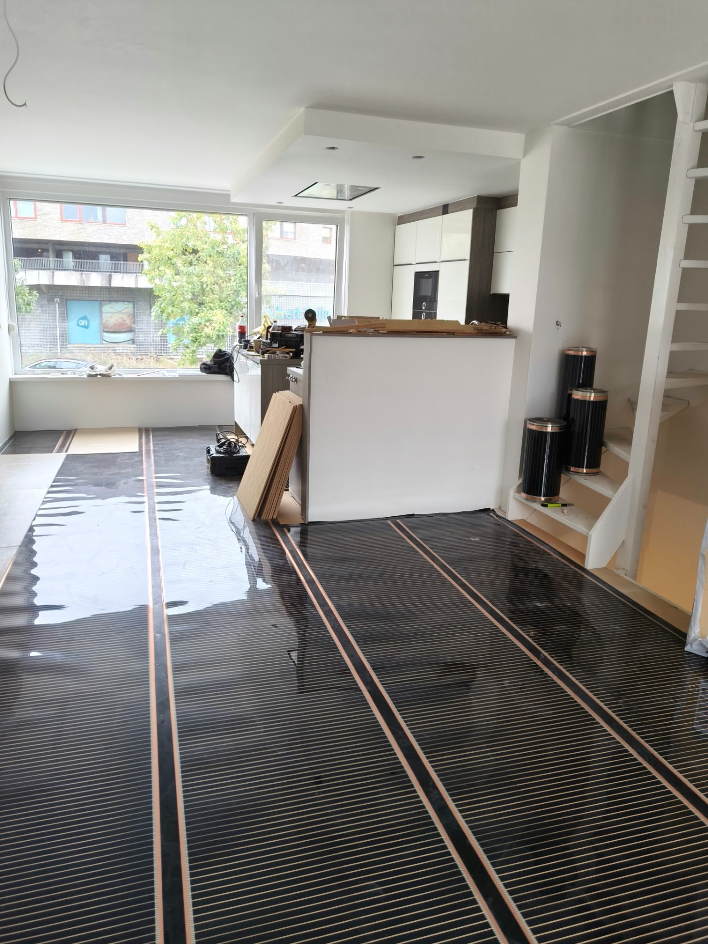 Woonkeuken in verbouwing met vloerverwarming, grote ramen, keukeneiland en opgestapelde materialen.