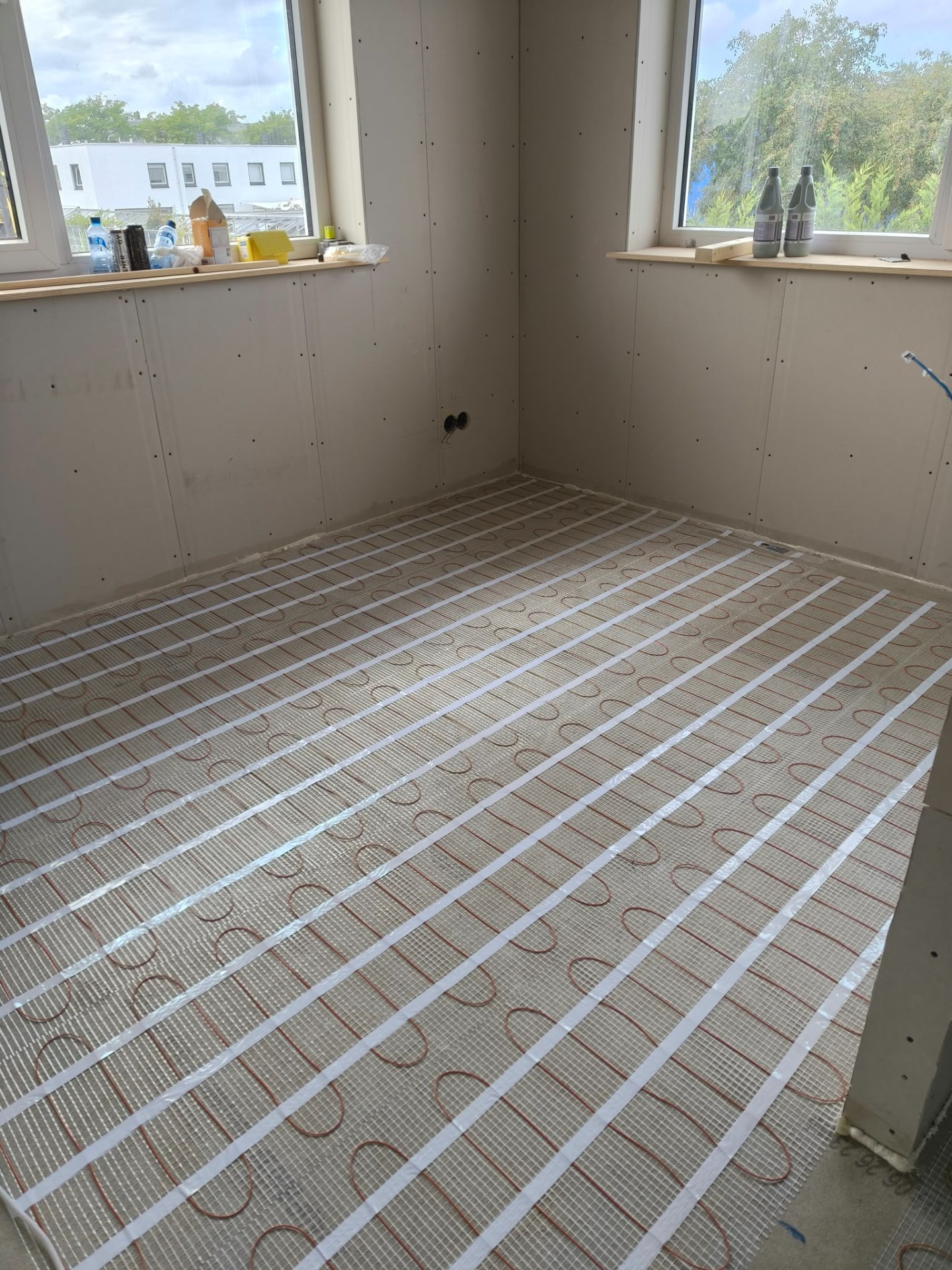 Vloerverwarming met kabels op rastermatten in lege ruimte, omringd door gipsplaten en ramen.