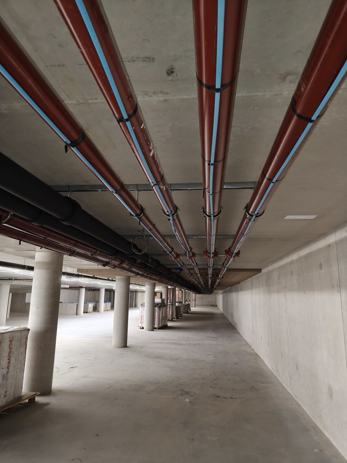 Rij metalen leidingen met blauwe elektrische tracing kabel langs betonnen plafond, in lege parkeergarage.