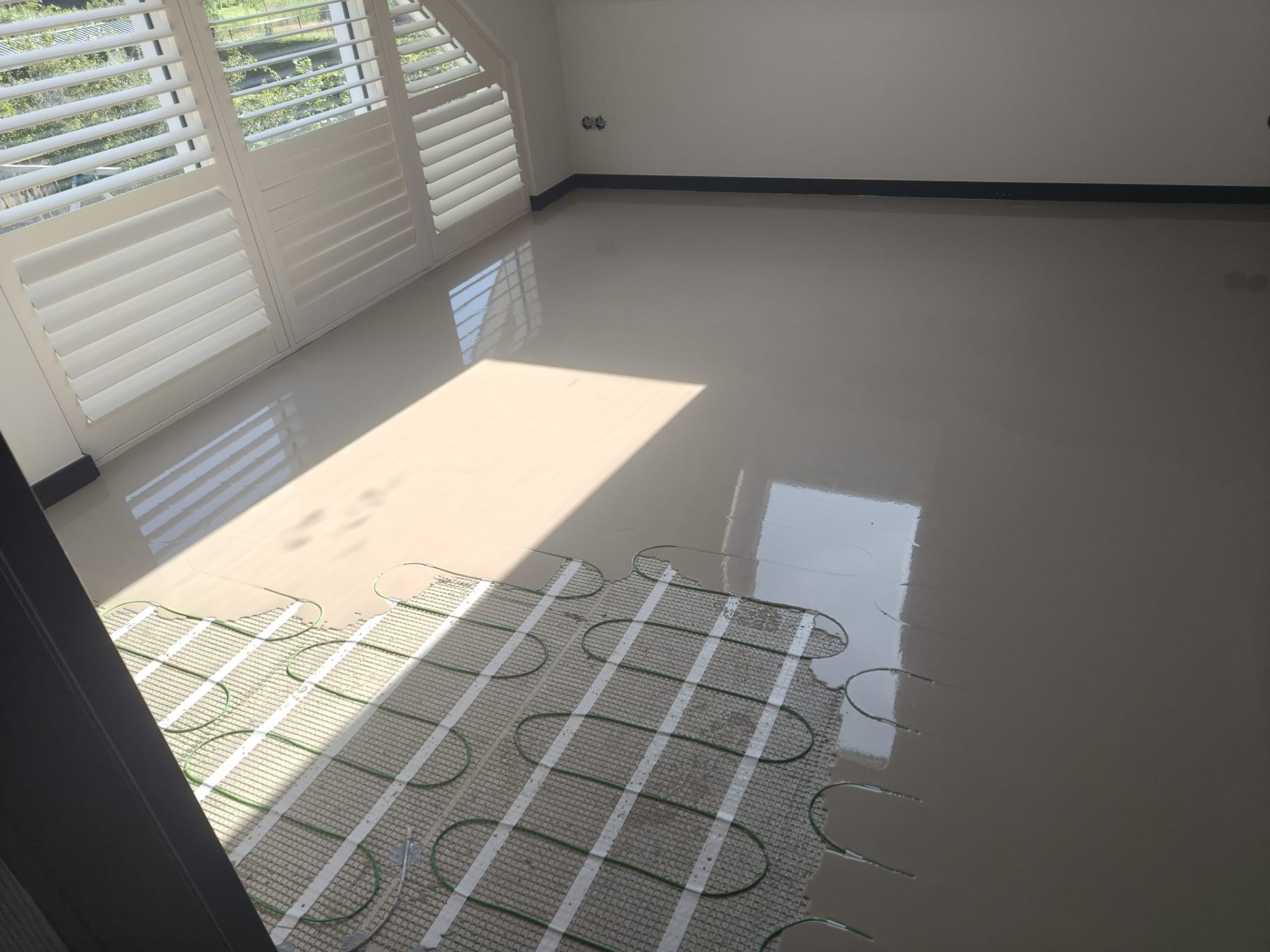 Lege kamer met witte shutters, grijze vloer en vloerverwarming met groene leidingen, zonlicht reflecteert.