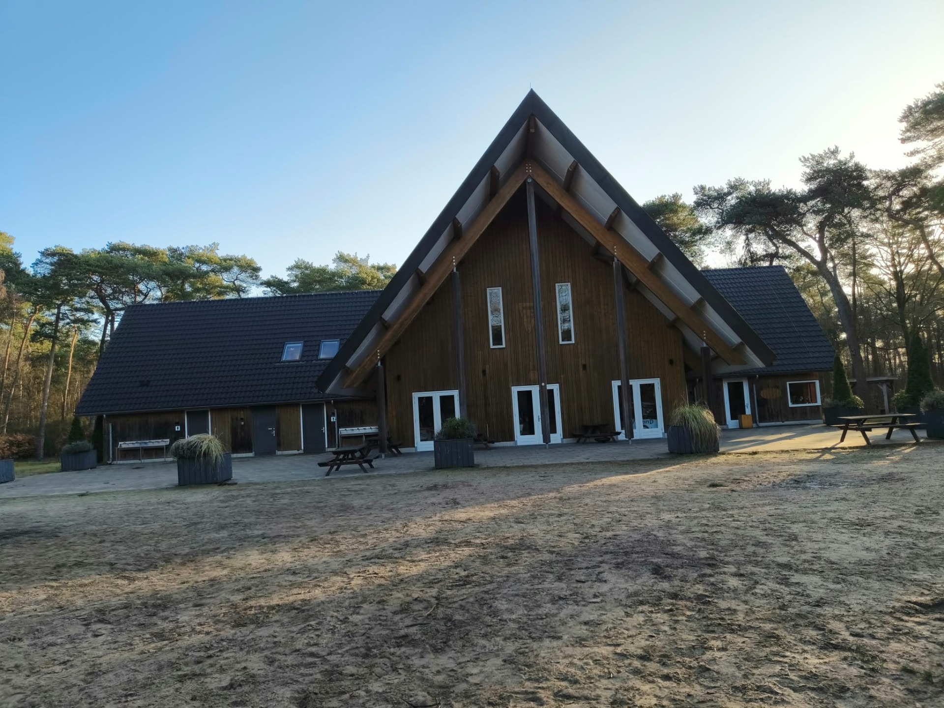Houten boerderij met puntgevel, zwarte dakpannen, glazen deuren en banken op zandpad, omringd door dennen.